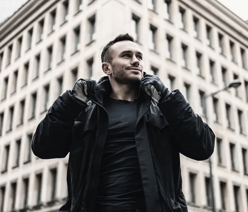 Maciej Łyszczarz w ciemnej kurtce na tle warszawskiego budynku